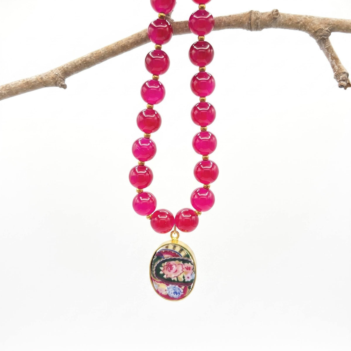 Burgundy and gold beaded necklace with rose porcelain pendant, hanging on a branch.