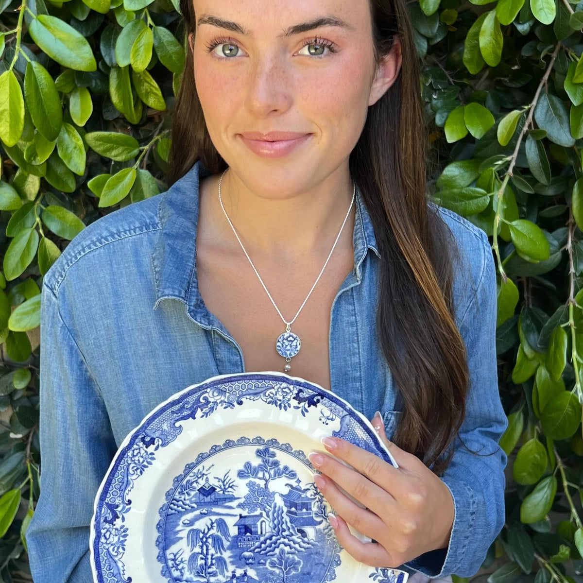 May include: A woman wearing a blue denim shirt holds a blue and white porcelain plate. The plate features a detailed landscape design. She also wears a silver necklace with a pendant that has a blue and white floral design.