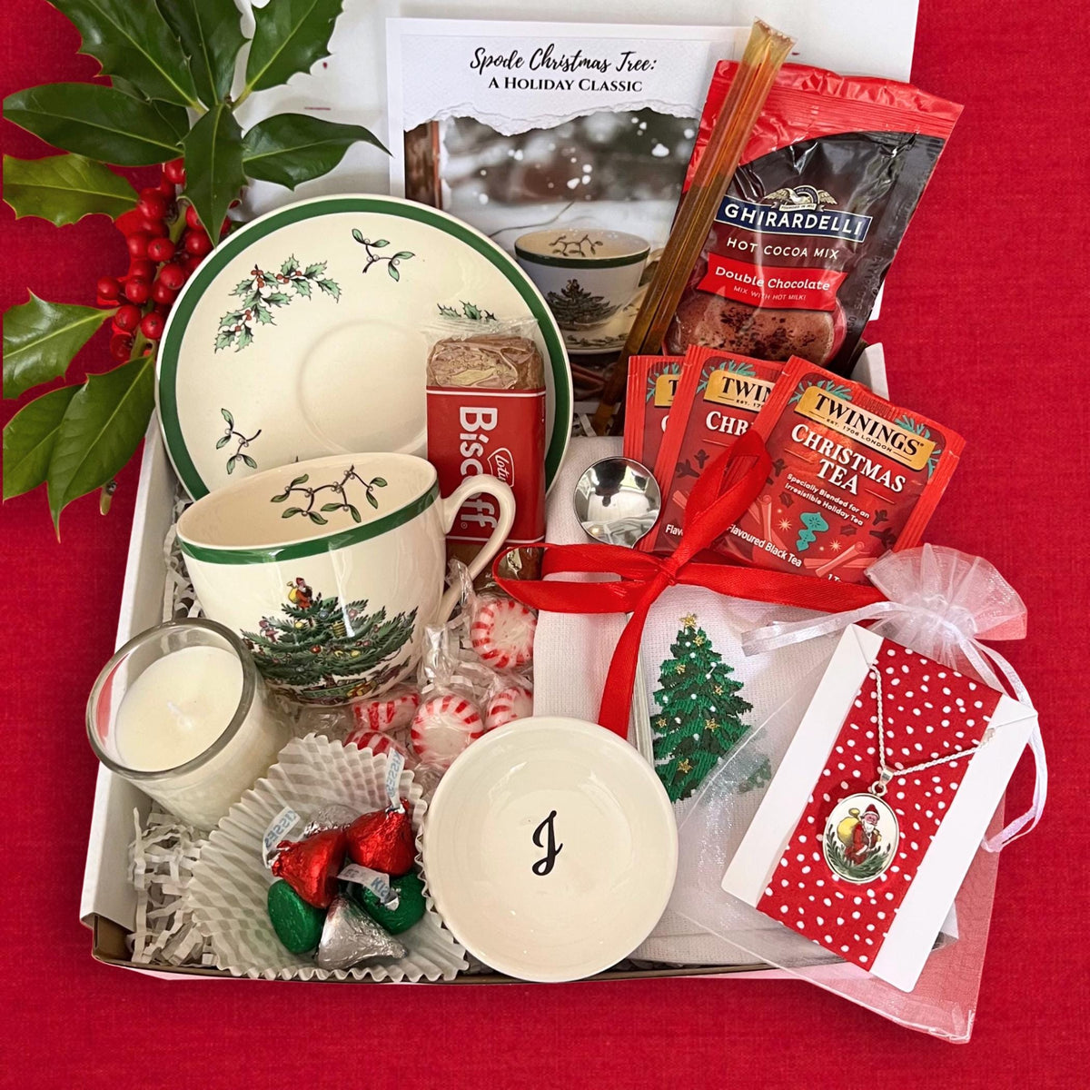 May include: A holiday gift box filled with a Spode Christmas Tree teacup and saucer, hot cocoa mix, tea bags, a small bowl with the letter "J", a candle, and a necklace. The box is tied with a red ribbon and includes candy.