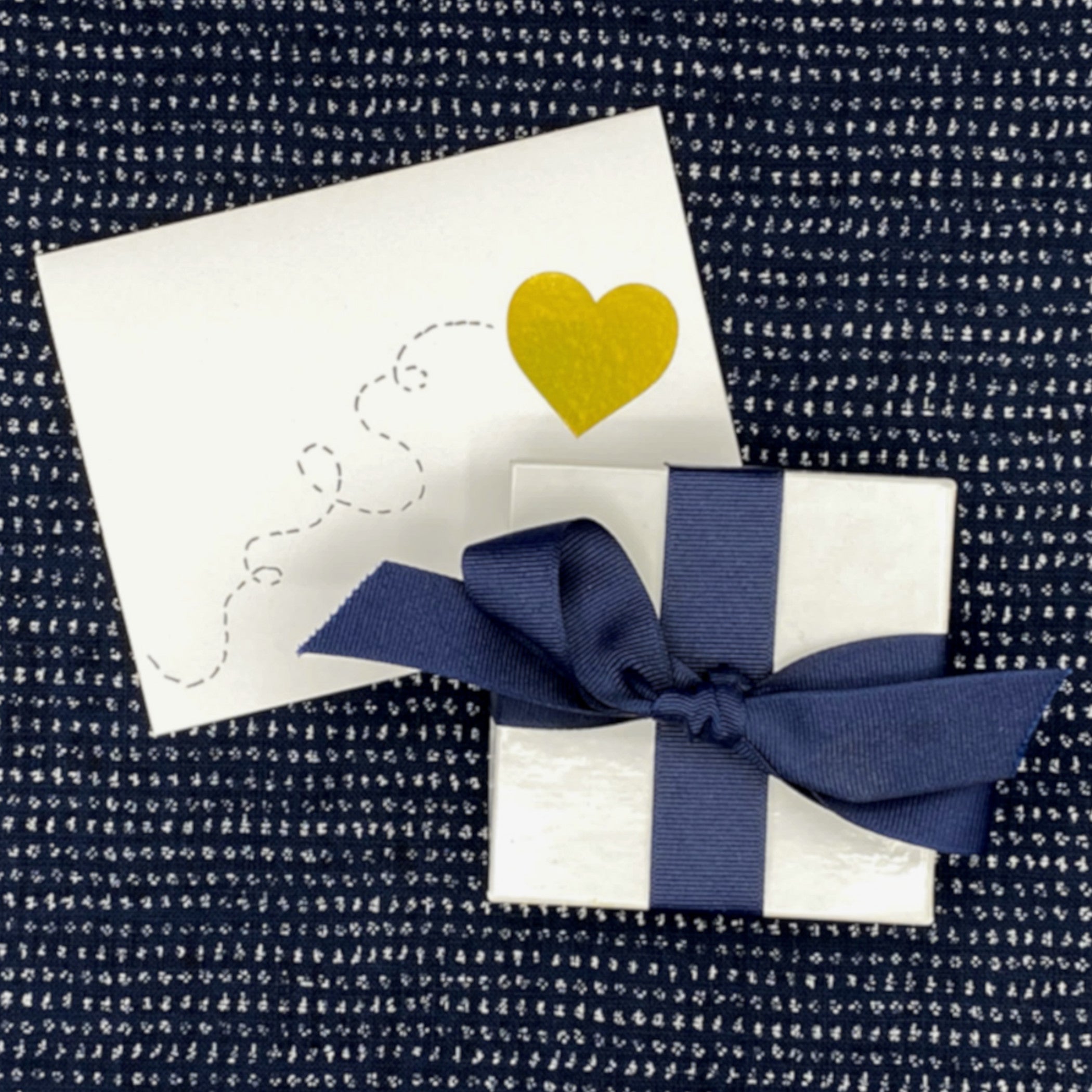 Gift box with navy ribbon and a card with a gold heart on textured fabric.