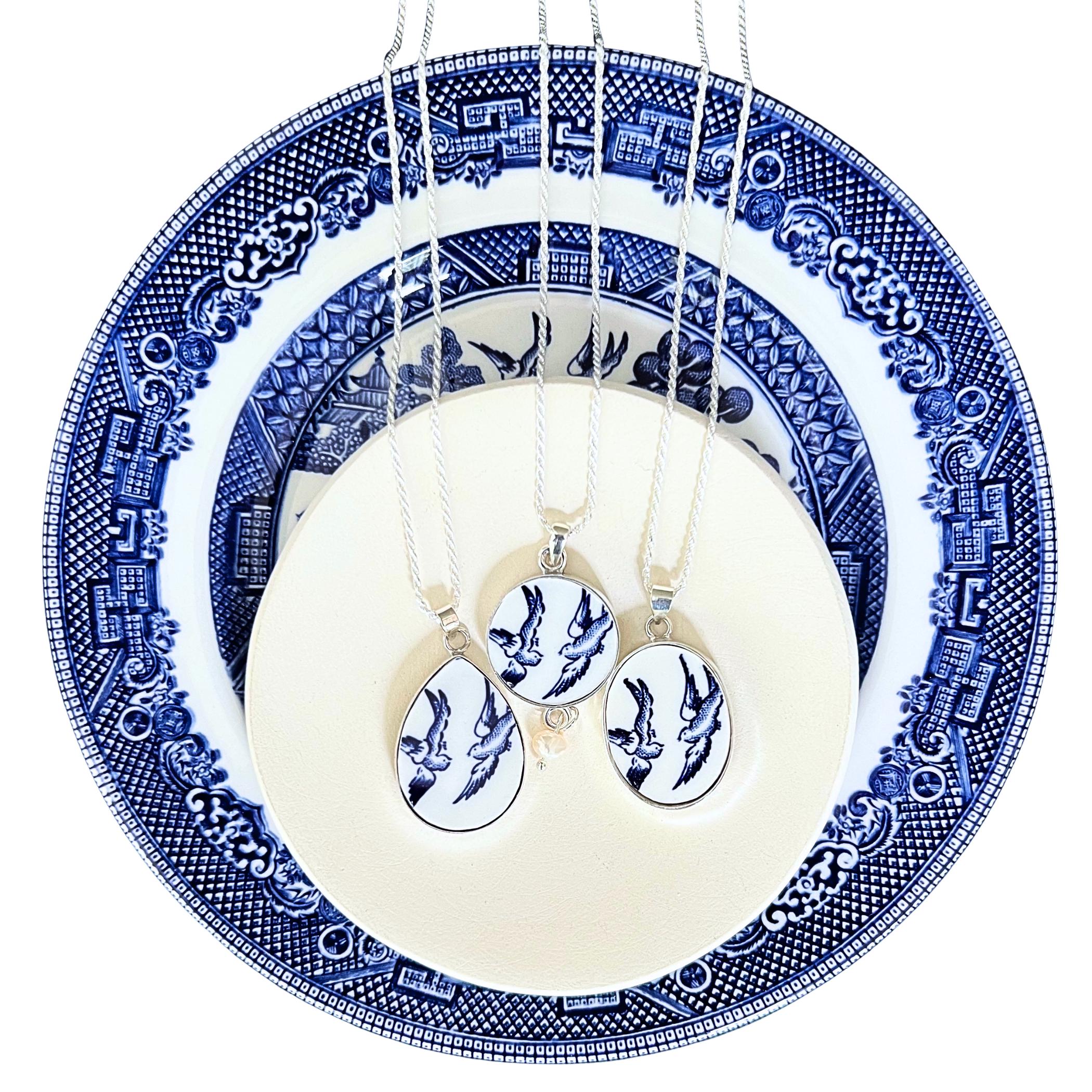 Three Blue Willow Love Birds necklaces — the oval, teardrop, and round pendants with chains — displayed on the vintage china plate they were cut from.