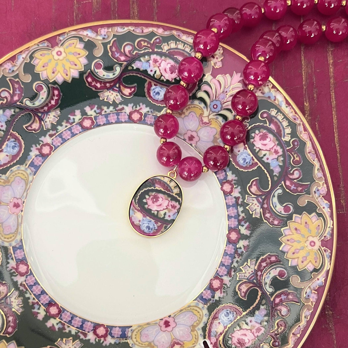 Burgundy and gold beaded necklace with a rose porcelain pendant, displayed on the antique Muirfield Royal Paisley plate it was upcycled from.